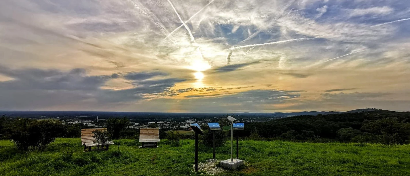 panorama_grosser_berg.jpg Sonnenuntergang über einer grünen Landschaft mit Info-Tafeln und Parkbänken unter dramatischem Himmel.