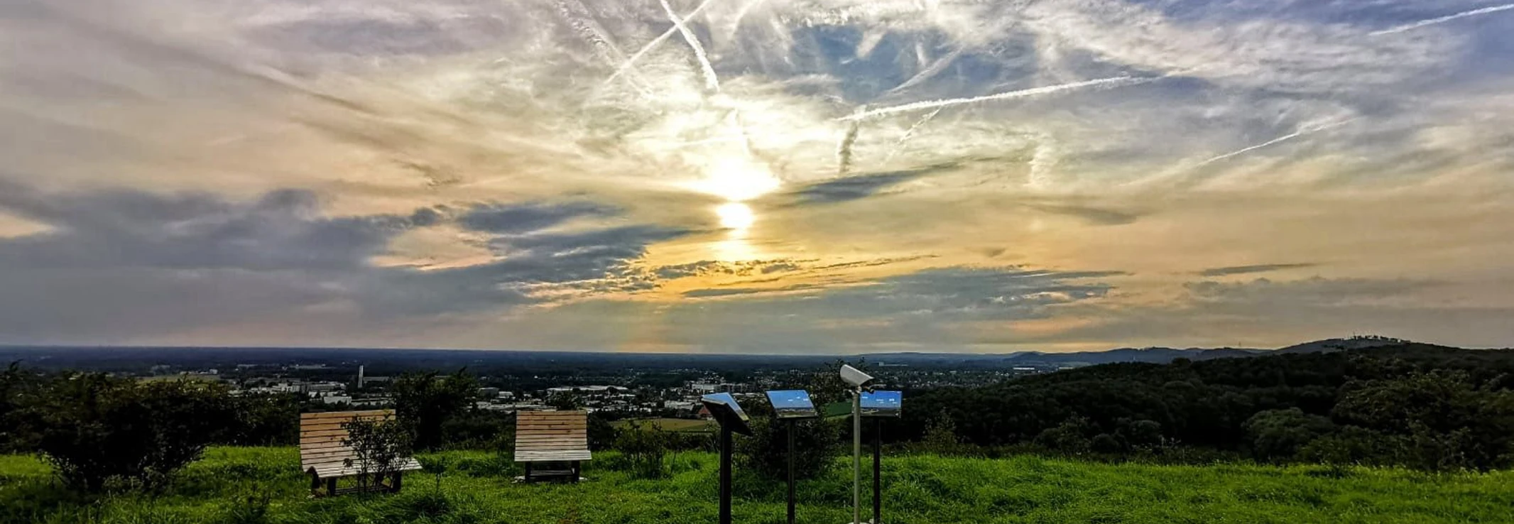 panorama_grosser_berg.jpg Sonnenuntergang über einer grünen Landschaft mit Info-Tafeln und Parkbänken unter dramatischem Himmel.