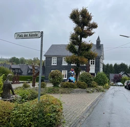 Kunst Kabinett Hesper Der Platz der Künste in Reichshof zeigt Skulpturen umgeben von Gebäuden und herbstlichen Bäumen.