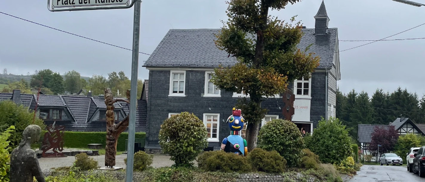 Kunst Kabinett Hesper Der Platz der Künste in Reichshof zeigt Skulpturen umgeben von Gebäuden und herbstlichen Bäumen.
