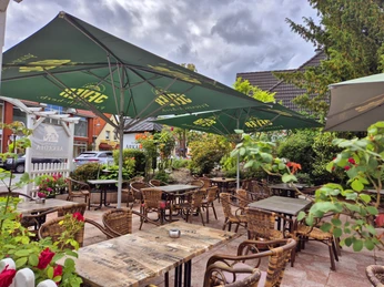 Außenansicht Restaurant Arkadia Ein gemütlicher Außenbereich eines Cafés mit rustikalen Tischen und Stühlen, umgeben von Grünen Pflanzen. Große Sonnenschirme spenden Schatten an einem wolkigen Tag.