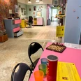 Museum des Schmerzes und der Liebe Im Vordergrund steht ein rosafarbener Tisch mit bunten Bechern, einer Chipstube und einem Schachbrett.In the foreground, there is a pink table with colorful cups, a tube of chips, and a chessboard.