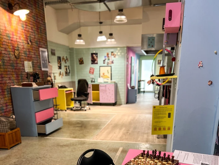 Museum des Schmerzes und der Liebe Im Vordergrund steht ein rosafarbener Tisch mit bunten Bechern, einer Chipstube und einem Schachbrett.In the foreground, there is a pink table with colorful cups, a tube of chips, and a chessboard.