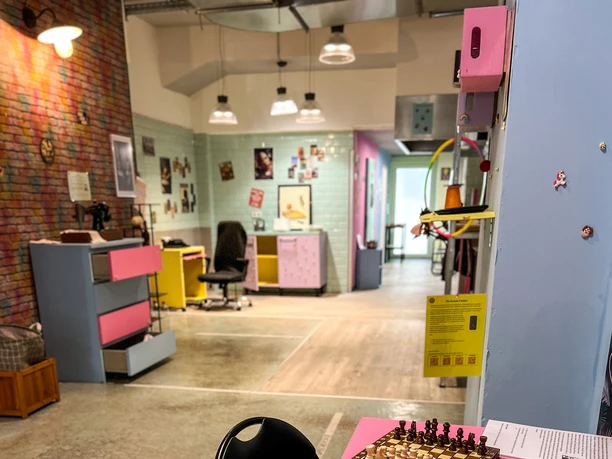 Museum of pain and love In the foreground, there is a pink table with colorful cups, a tube of chips, and a chessboard.