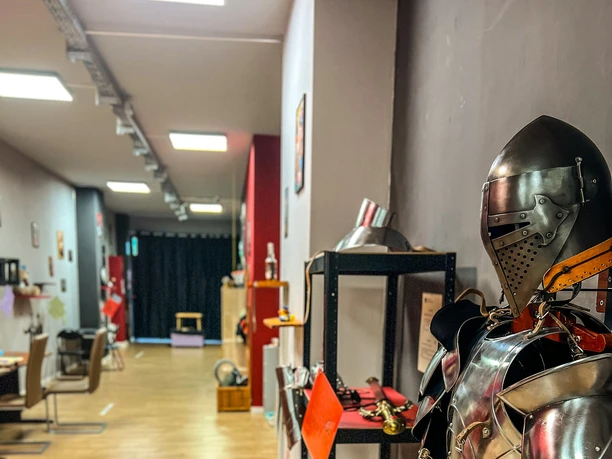 Museum of pain and love The image shows a detailed knight's armor made of shiny metal, displayed in a modernly designed museum space. In the background, various exhibits and furniture are visible on a light wooden floor.