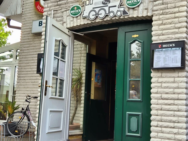 BaB Bistro am Bahnhof Vorderansicht Vorderansicht einer gemauerten Fassade mit grünen Türen und offenem Torbogen unter einem Bierwerbeschild. Ein Fahrrad steht links vom Eingang, signalisiert ein lokal etabliertes Ambiente.