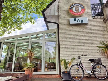 BaB Bistro am Bahnhof Außenfassade Das Bild zeigt die Fassade eines gemütlichen Restaurants mit einer Veranda, dekoriert mit grünen Pflanzen und einem markanten Schild der Marke Erdinger Weißbräu, unter blauem Himmel.