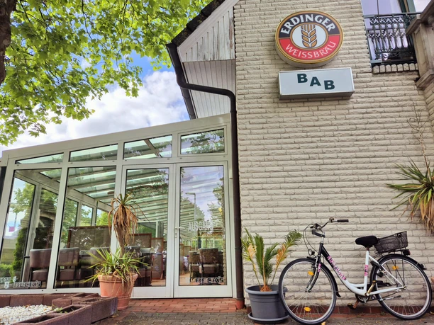BaB Bistro am Bahnhof Außenfassade Das Bild zeigt die Fassade eines gemütlichen Restaurants mit einer Veranda, dekoriert mit grünen Pflanzen und einem markanten Schild der Marke Erdinger Weißbräu, unter blauem Himmel.