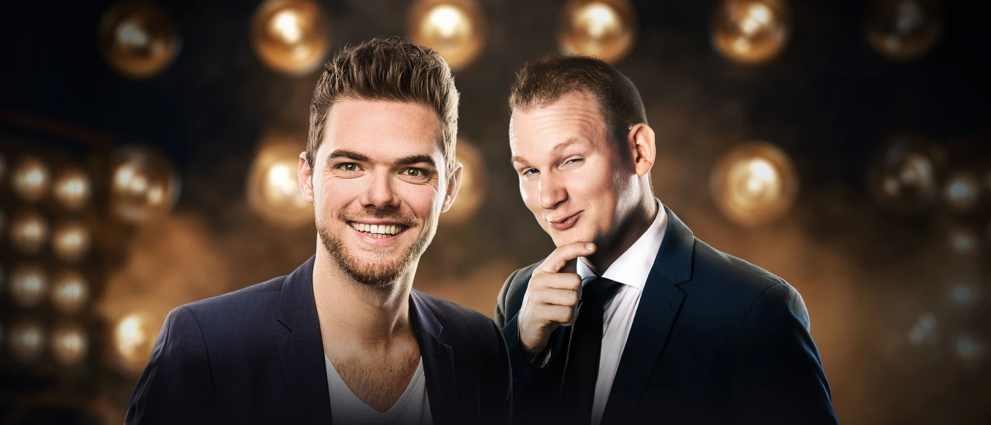 Zwei Männer vor Scheinwerfer-Hintergrund, die Anzüge tragen und lachend in die Kamera schauen.