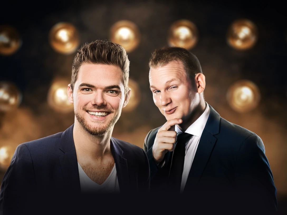 Die Unfassbaren (c) Niclas Mettendorf.jpg Zwei Männer vor Scheinwerfer-Hintergrund, die Anzüge tragen und lachend in die Kamera schauen.