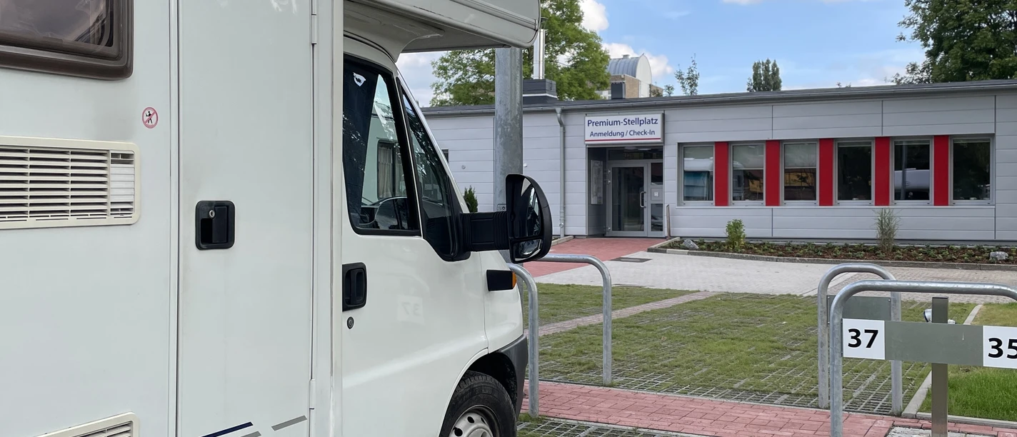 ```html Ein Wohnmobil parkt auf einem Rasengitterplatz vor einem modernen Gebäude mit großen Fenstern. ```
