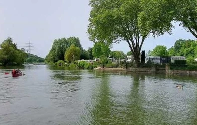 Bild zeigt einen Fluss mit Boot, umgeben von Bäumen und einem Wohnmobilplatz am Ufer.