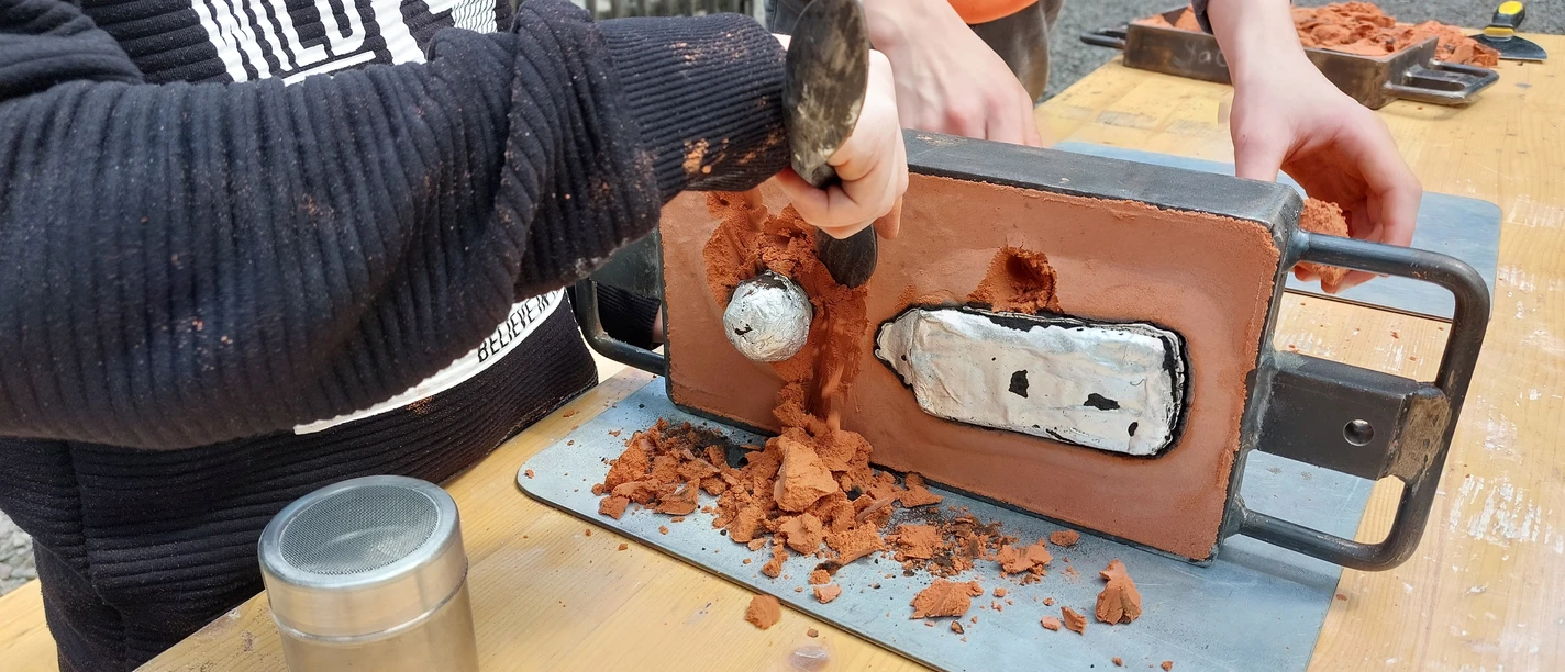 Metallgießen am Oelchenshammer Zwei Personen arbeiten an einem metallgerahmten Tonmodell und formen mit Werkzeugen feine Details heraus.