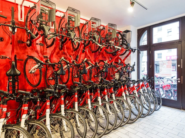 Colonia Aktiv Fahrradtouren in Köln Ein heller Raum mit roten Wänden, in dem rote Fahrräder an der Wand hängen und in Reihen stehen.