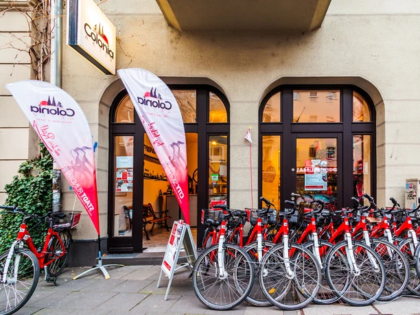 Colonia Aktiv Fahrradtouren in Köln Vor dem Eingang von Colonia Aktiv stehen zahlreiche rote Fahrräder geordnet in einer Reihe. Zwei weiße Fahnen mit dem Colonia-Aktiv-Logo sind sichtbar. Der Hintergrund zeigt große Fenster und einen Eingang zu einem Gebäude.