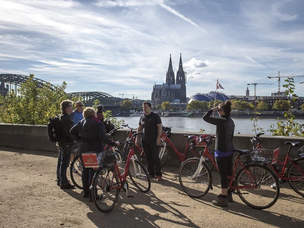 Colonia Aktiv guided bike tours in Cologne A group of people stands with bicycles against the backdrop of Cologne Cathedral on the banks of the Rhine. The atmosphere is relaxed, under sunny conditions.