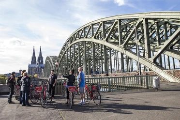 Colonia Aktiv Fahrradtouren in Köln Gruppe von Menschen mit roten Fahrrädern auf einer Brücke in Köln, Dom im Hintergrund.