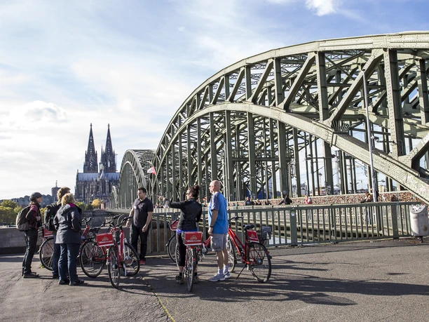 Colonia Aktiv guided bike tours in Cologne Group of people with red bicycles on a bridge in Cologne, with the Cologne Cathedral visible in the background.