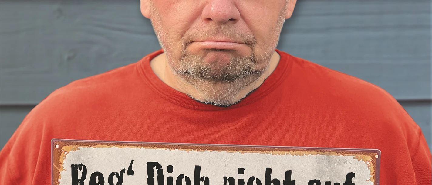 Bernd_Stelter_RegDichnichtauf_Credits_Stelter_13x18.jpg Mann mit Brille in rotem Pulli hält Schild mit humorvoller Botschaft gegen Sorgen hoch.