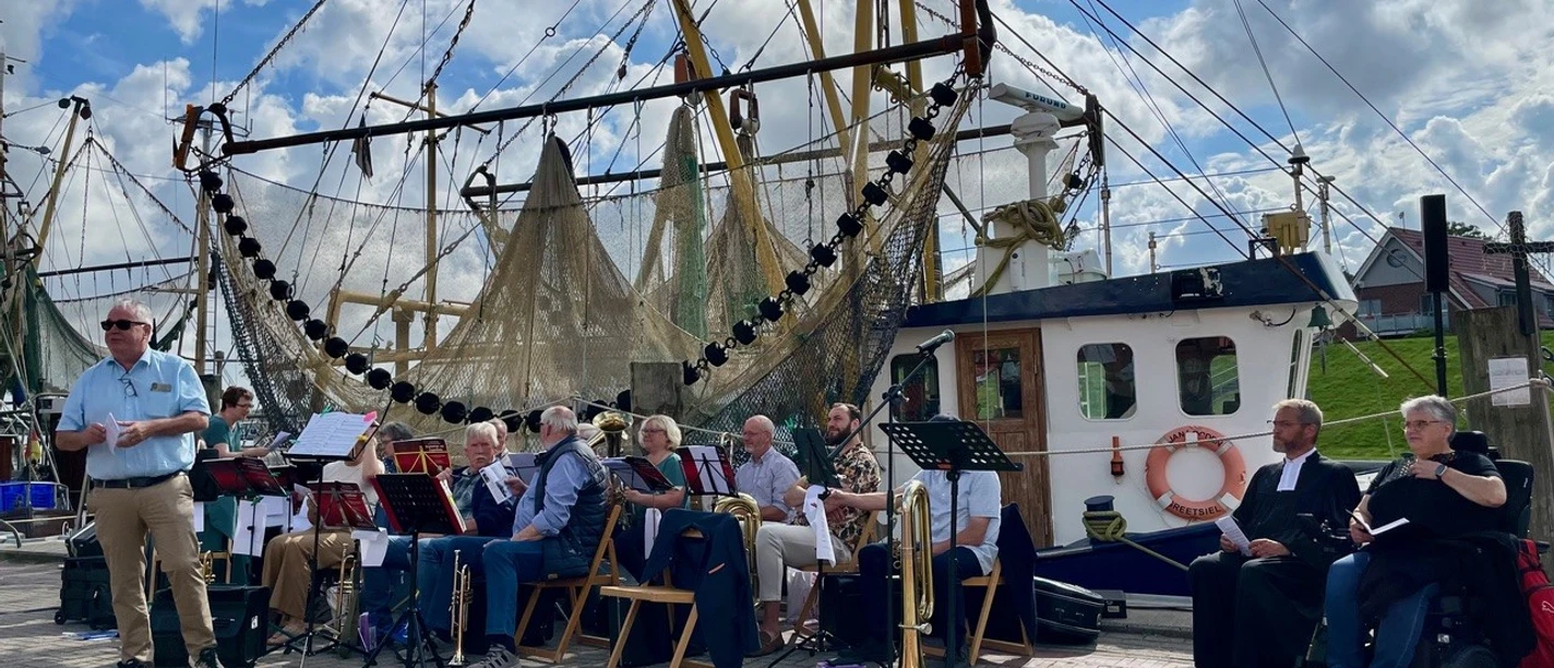 Hafengottesdienst Blick auf einen Kutter der im Hafen Greetsiel liegt. Davor sitzen Mitglieder vom Posaunenchor.