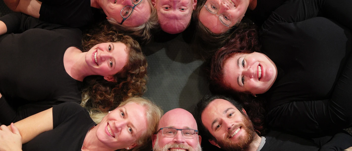 Improthesen_Foto_Klawunn_4687a (Flyerfoto).jpg Eine Gruppe von acht Menschen liegt lächelnd in einem Kreis auf dem Boden und blickt zur Kamera.