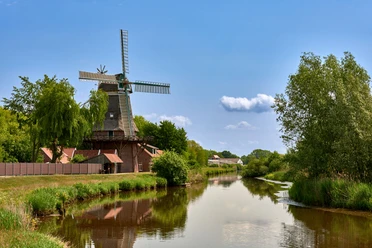 Hengstforder Mühle_Foto Ostfriesland Touristik.jpg Historische Windmühle am Flussufer mit blauer Himmelskulisse und üppigem Grün