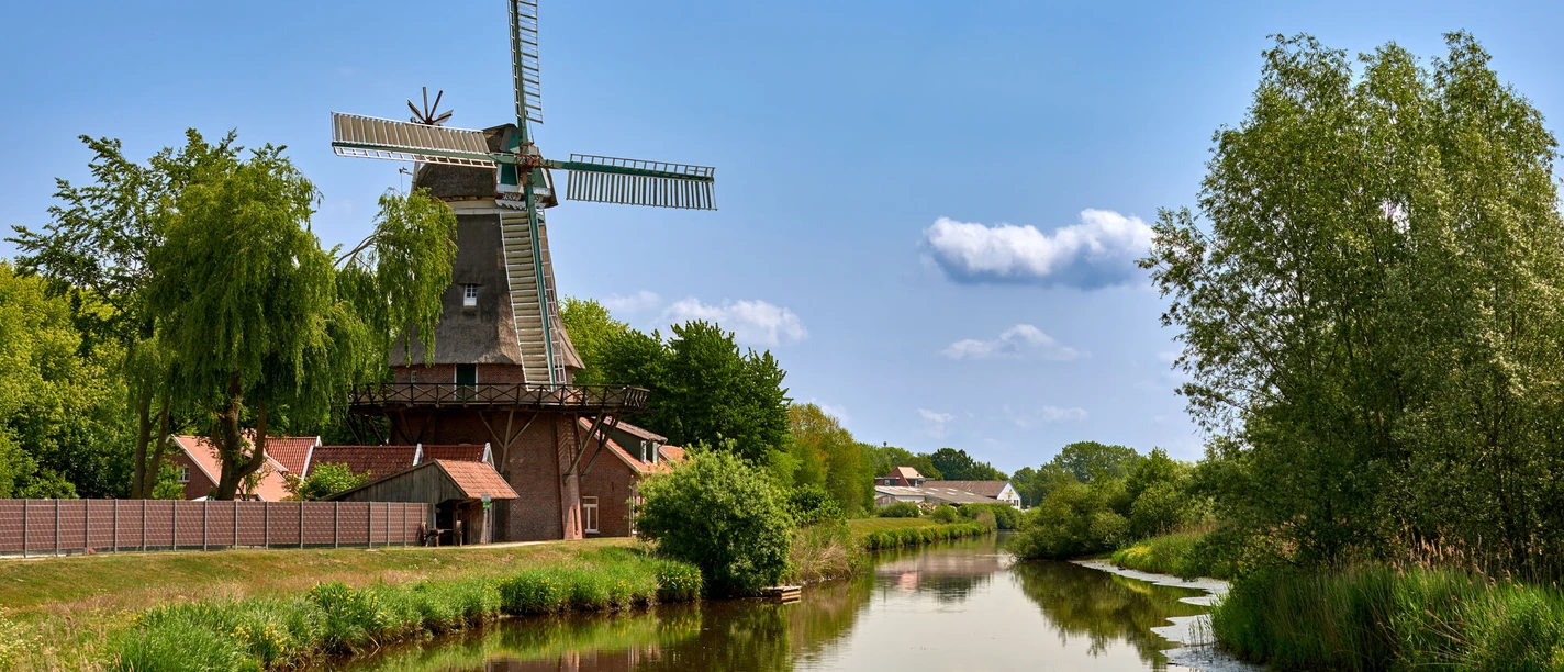 Hengstforder Mühle_Foto Ostfriesland Touristik.jpg Historische Windmühle am Flussufer mit blauer Himmelskulisse und üppigem Grün