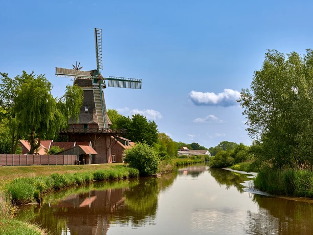 Hengstforder Mühle_Foto Ostfriesland Touristik.jpg Historische Windmühle am Flussufer mit blauer Himmelskulisse und üppigem Grün