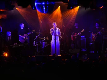 Club Bahnhof Ehrenfeld Ein Musiker steht auf der Bühne, umgeben von einer Band, die in schummriges Licht getaucht ist.A musician stands on stage, surrounded by a band bathed in dim light.