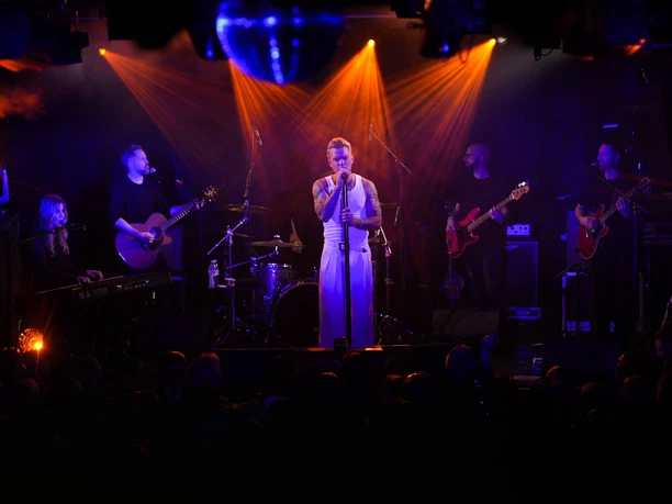 Club Bahnhof Ehrenfeld A musician stands on stage, surrounded by a band bathed in dim light.
