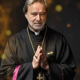 Kardianalfehler, Neues Theater Espelkamp Das Bild zeigt einen älteren Mann mit grauem Haar und Bart in traditioneller Kleidung eines katholischen Geistlichen. Er trägt ein schwarzes Gewand mit rosafarbenen Akzenten und ein goldfarbenes Kreuz um den Hals. Seine Hände sind zum Gebet gefaltet.
