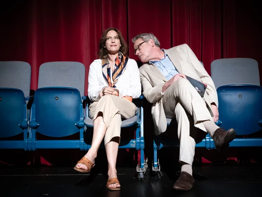 Was war und was wird, Neues Theater Espelkamp In einem modernen Theaterambiente sitzen eine Frau und ein Mann in eleganter Kleidung auf blauen Polstersesseln. Eine rote Bühne ist im Hintergrund zu sehen.