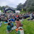 Auf einer Wiese in einem Park sitzen viele Menschen und Familien mit dem Blick auf eine Open-Air-Bühne gerichtet, auf der ein Theaterstück aufgeführt wird.