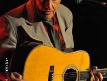 Ein Musiker mit grauem Anzug und Hut spielt eine akustische Gitarre auf einer Bühne vor dunklem Hintergrund.