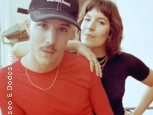 Zwei lächelnde Personen in legerer Kleidung posieren vor einer urbanen Kulisse in Nahaufnahme.