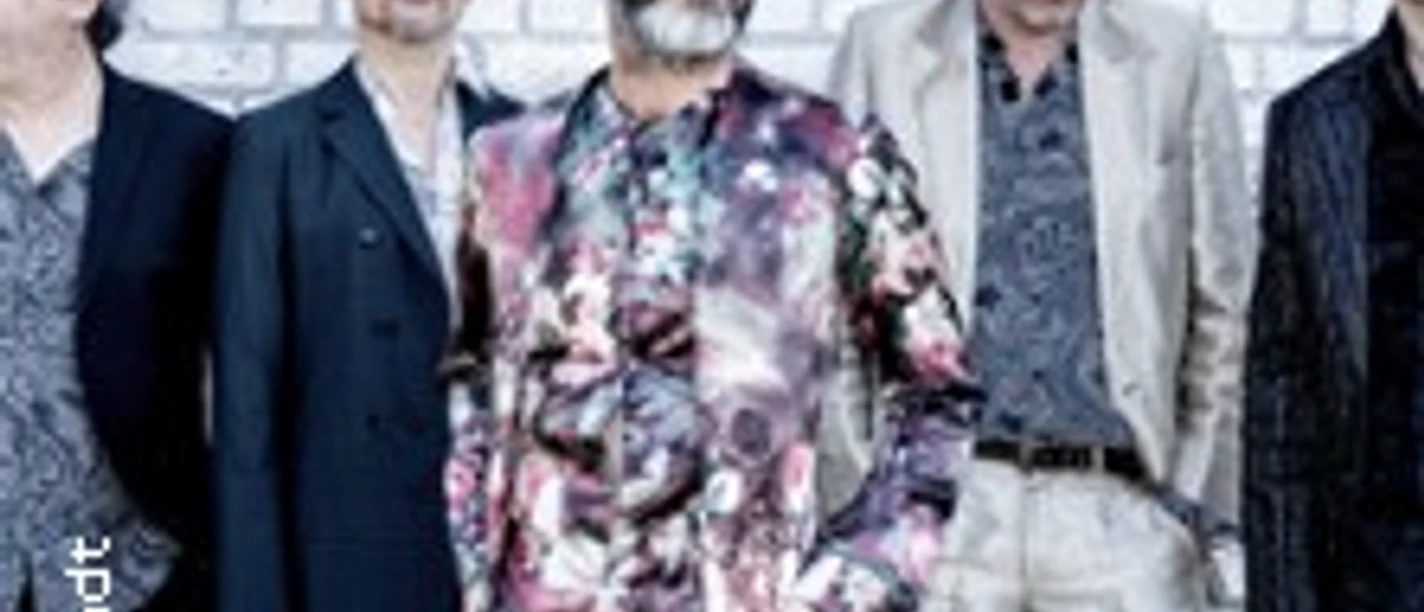 Fünf Männer in modischer Kleidung vor einer Graffiti-Wand, diverse Posen einnehmend.