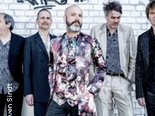 Fünf Männer in modischer Kleidung vor einer Graffiti-Wand, diverse Posen einnehmend.