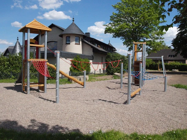 Spielplatz mit modernen Klettergerüsten aus Holz und Seilnetz, umgeben von Wohnhäusern und Bäumen.