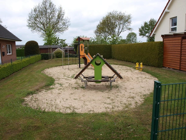Kleiner Spielplatz mit Kletterstruktur und Sandfläche, umgeben von Hecken und Häusern in ruhiger Lage.