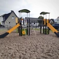 Zwei Rutschen und ein Klettergerüst aus Holz stehen auf dem Kiesboden eines Spielplatzes in Wohngebiet.