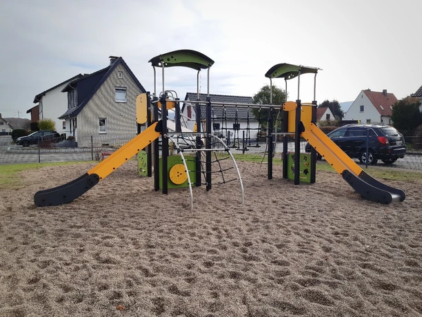 Zwei Rutschen und ein Klettergerüst aus Holz stehen auf dem Kiesboden eines Spielplatzes in Wohngebiet.