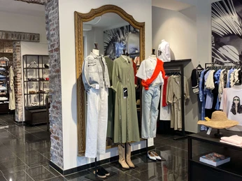 Simon und Renoldi Das Bild zeigt eine Boutique mit modernem Interieur. Zwei Schaufensterpuppen präsentieren modische Kleidung: ein khakifarbenes Kleid und ein Ensemble aus heller Bluse mit rotem Akzent und Jeans.The picture shows a boutique with a modern interior. Two mannequins present fashionable clothing: a khaki dress and an ensemble consisting of a light-colored blouse with a red accent and jeans.