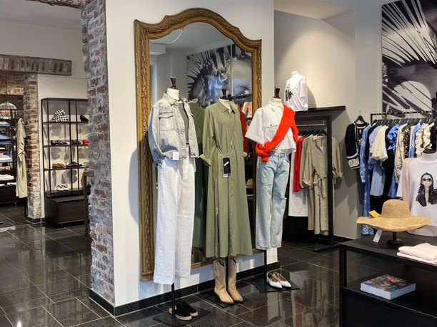 Simon und Renoldi Das Bild zeigt eine Boutique mit modernem Interieur. Zwei Schaufensterpuppen präsentieren modische Kleidung: ein khakifarbenes Kleid und ein Ensemble aus heller Bluse mit rotem Akzent und Jeans.