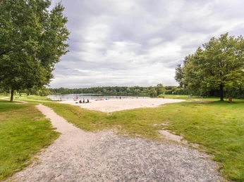 woldsee-badestrand-eingang.jpg Ein breiter, grasbewachsener Pfad führt zu einem sandigen Badestrand am idyllischen Woldsee, eingefasst von Bäumen.