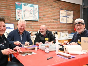 repair-cafe-bad-zwischenahn-messer-schleifen.jpg Vier lachende Personen sitzen an einem Tisch im Repair-Café und schleifen Messer. Ein Schild zeigt "Messer schleifen".