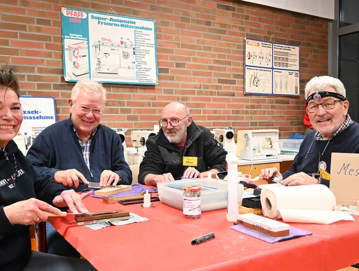 repair-cafe-bad-zwischenahn-messer-schleifen.jpg Vier lachende Personen sitzen an einem Tisch im Repair-Café und schleifen Messer. Ein Schild zeigt "Messer schleifen".