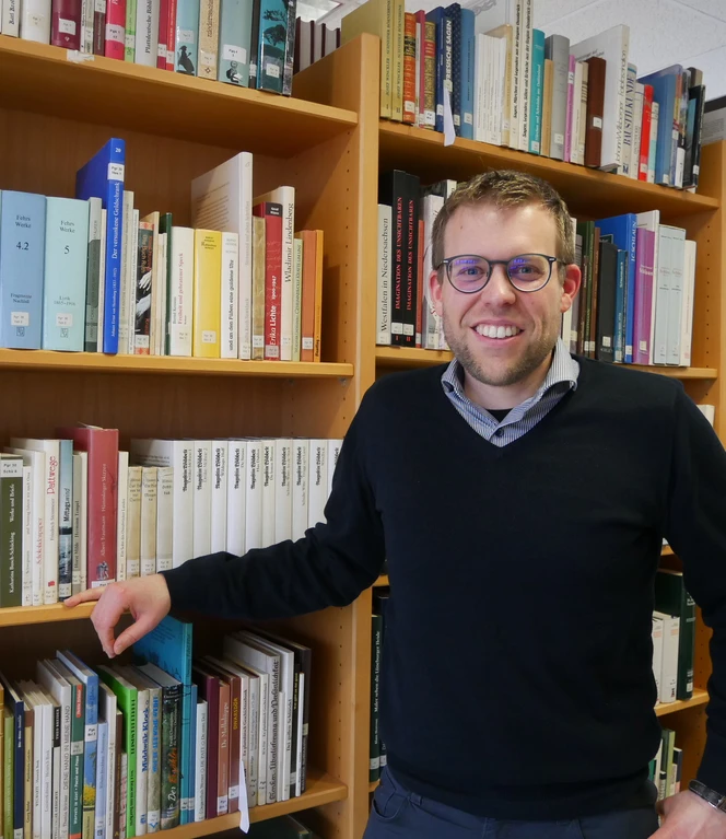 Dr. Benjamin van der Linde, Fachstelle Regionalgeschichte – Emsländischer Heimatbund © Christiane Adam Mann mit Brille steht lächelnd vor Bücherregalen in einer Bibliothek oder Facharchivumgebung.