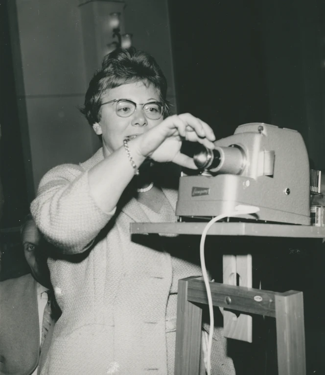 Elisabeth Schlicht, deren Wirken in der Ausstellung „Neustart Heimat. Dr. Elisabeth Schlicht und der Emslandplan“ thematisiert wird, bei einem ihrer zahlreichen Vorträge. Frau hält während eines Vortrags einen Diaprojektor und blickt konzentriert auf die Leinwand.