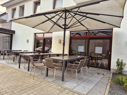 Bäckerei Schultze Mühlenfeld Außenansicht Das Bild zeigt eine moderne Terrasse eines Restaurants mit stilvollen Metall- und Kunststoffstühlen, Tischen und einem großen Sonnenschirm.