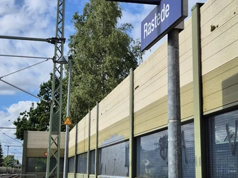 Bahnhof Rastede Bahnhof Rastede mit Gleisen, Schallschutzwand und Schild Richtung Wilhelmshaven bei bewölktem Himmel.